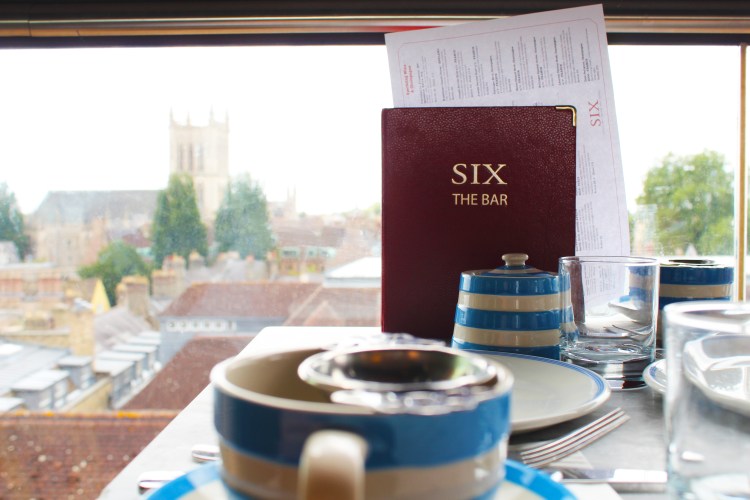 Close up of blue and white striped tea set with a red menu leaning against a window with a background view of Cambridge city.