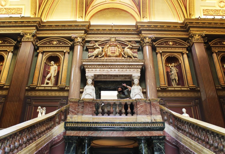 Top of the staircase with a grand entrance surrounded by sculptures and historical art with a brunette women posing at the top.